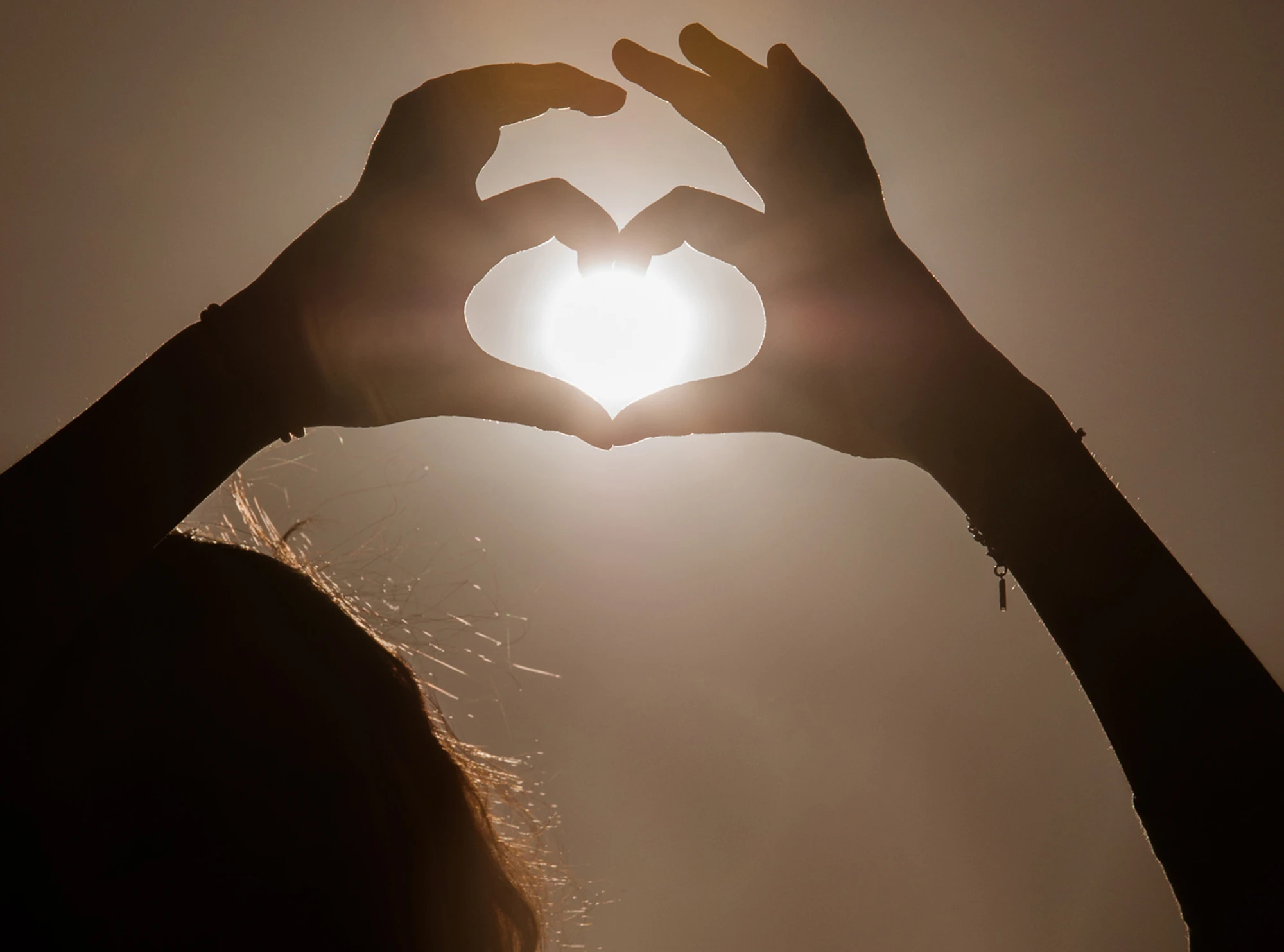 Person bildet mit Händen Herz um Sonne am Himmel, in Brauntönen gehalten