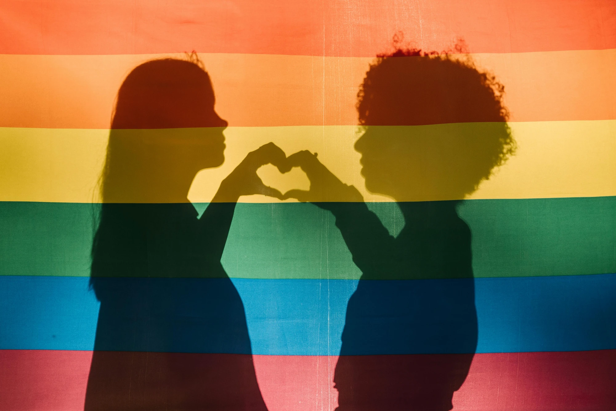Regenbogenflagge, dahinter als Schattenfiguren ein Paar, das mit den einander berührenden Fingern ein Herz formt